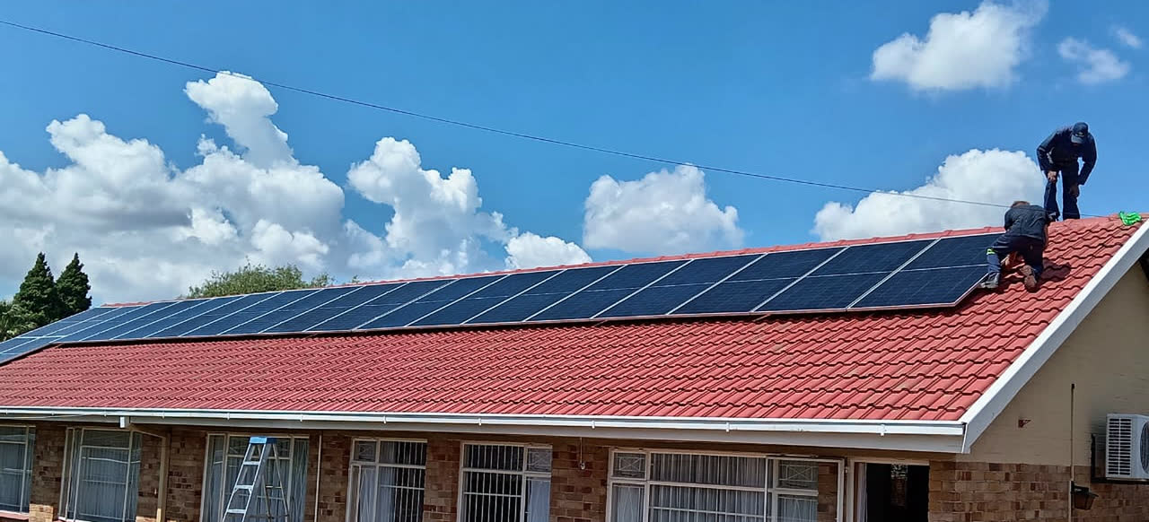 A technician standing on a tiled roof beside a ladder, installing a solar panel array under a clear sky